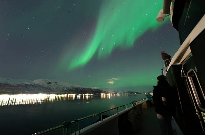 View from a boat deck of green aurora over a fjord with city lights on the shore