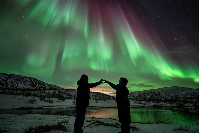 Two silhouettes making a heart shape with their hands under green aurora