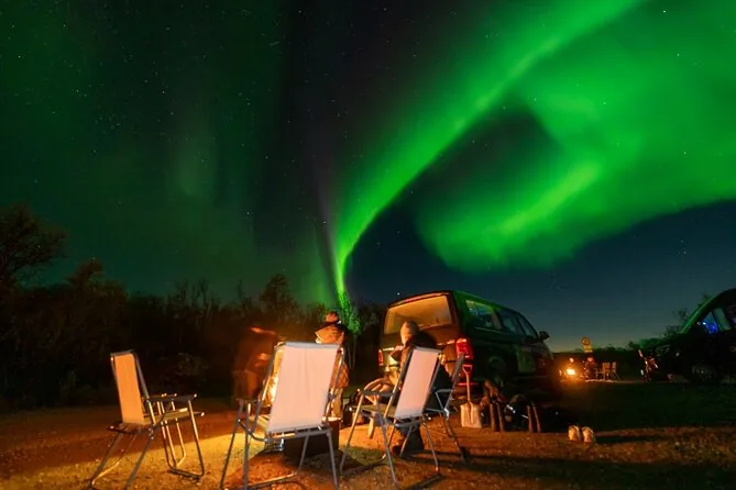 Sillas de camping junto a una furgoneta y una hoguera bajo una aurora verde